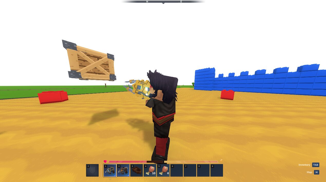 A player aims the physgun at a wooden crate held in the air, with a blue brick wall and a red block on the sand.